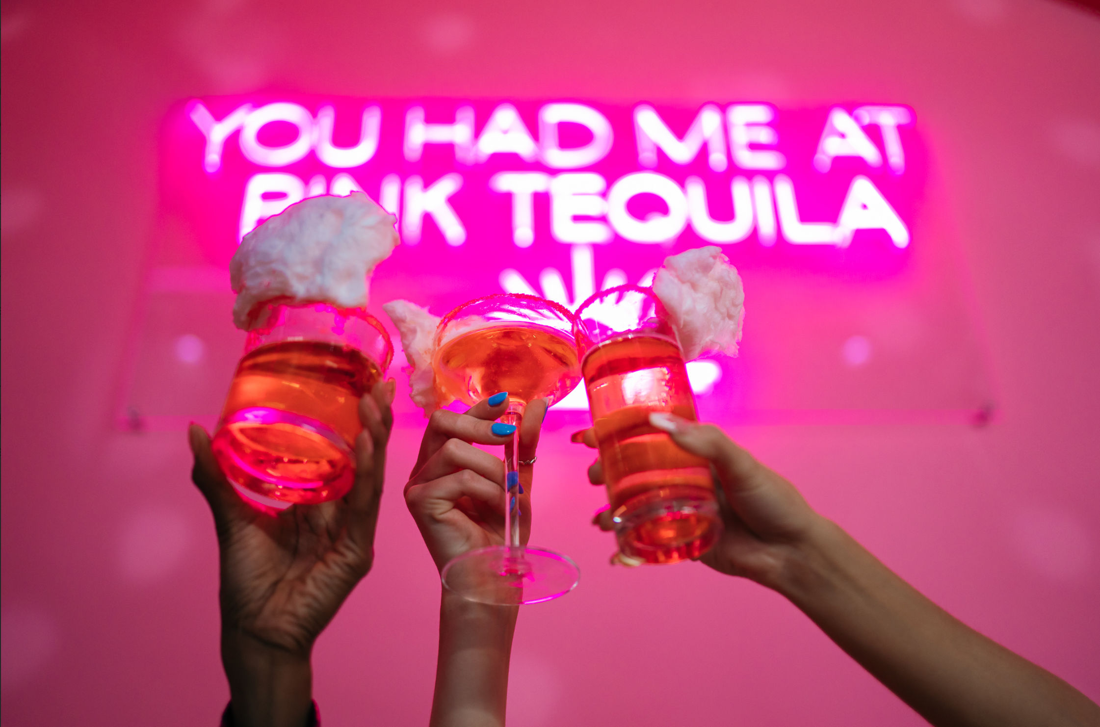 Three hands toasting pink cocktails topped with cotton candy beneath a neon sign that reads "YOU HAD ME AT PINK TEQUILA" in a trendy pink cocktail bar, playful nightlife vibe.