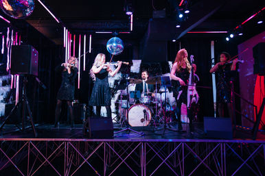 String quartet with drummer performing on a neon-lit nightclub stage under a disco ball, musicians in sparkling black dresses.