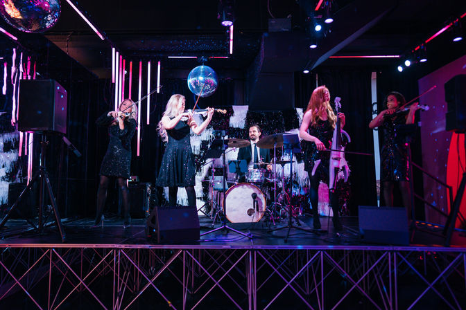 String quartet with drummer performing on a neon-lit nightclub stage under a disco ball, musicians in sparkling black dresses.