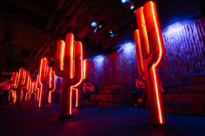 Indoor neon cactus art installation with tall red-orange LED-outlined cactus sculptures, blue-purple ambient lighting, tiered seating and visitors in the background