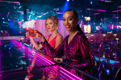 Two glamorous women in sequined dresses toasting cocktails topped with cotton candy at a neon-lit city nightclub bar, vibrant pink and blue lights reflecting on a mirrored counter.