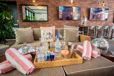 Stylish indoor lounge with brick wall and cushioned sofa, wicker tray with a vodka bottle, cocktail glass, ice bucket and glassware, pink-and-white striped towels, mirrored disco balls, TV screens and bar stools in the background