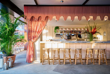 Sunlit poolside resort bar with red-and-white striped awning, marble countertop, six rattan bar stools, tropical potted palms, lanterns and a bright floral arrangement.