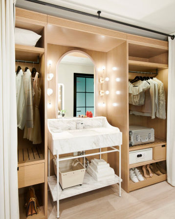 Chic modern walk-in closet dressing nook with marble vanity sink, arched lighted mirror, wood built-in wardrobe shelving with hanging neutral clothes, suitcase, folded towels and shoes.