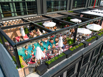 Aerial view of a lively rooftop pool party on a sunny afternoon — swimmers in a small urban hotel pool, sunbathers on red lounge chairs under white umbrellas, planters and a modern glass-and-brick building facade.