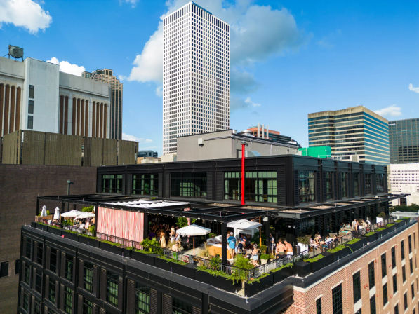 Vibrant rooftop bar and patio on a downtown building with umbrellas, greenery and people, set against a blue-sky city skyline.