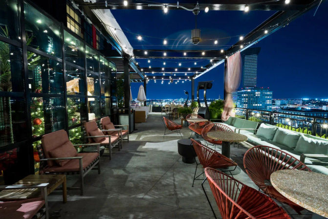 Rooftop terrace bar at night with string lights, cozy lounge sofas, red wire chairs and small tables overlooking an illuminated urban skyline.