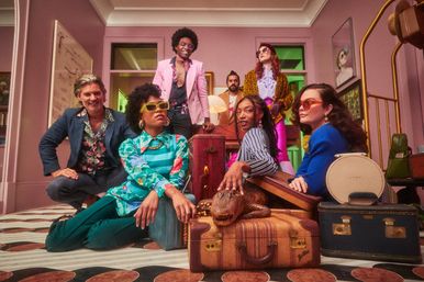 Stylish group portrait in a retro-inspired living room with colorful 1970s-style outfits and bold sunglasses, gathered around vintage suitcases and a decorative alligator prop.