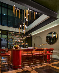 Glam retro‑chic hotel cocktail bar with red velvet stools and brass frames at a rounded red bar, backlit liquor shelves, globe pendant lights with gold fringe, round wall mirror and warm wood checkerboard floor — upscale city lounge vibe.