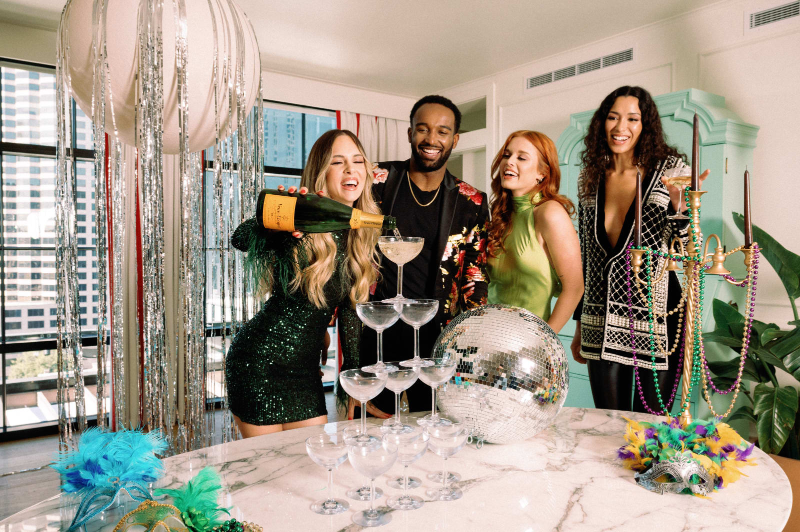 Four friends in a high-rise apartment celebrating with a champagne coupe tower, disco ball, feather masks, colorful beads and tinsel decorations