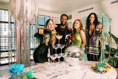 Four friends in a high-rise apartment celebrating with a champagne coupe tower, disco ball, feather masks, colorful beads and tinsel decorations