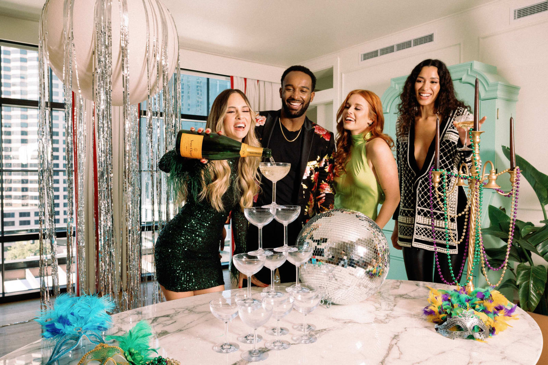 Four friends in a high-rise apartment celebrating with a champagne coupe tower, disco ball, feather masks, colorful beads and tinsel decorations