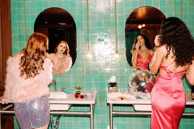 Two women getting ready at a retro green-tiled bathroom vanity: one in a pink feather jacket and sequin skirt touches up makeup in an oval mirror, the other in a satin pink dress applies lipstick beside a disco ball.