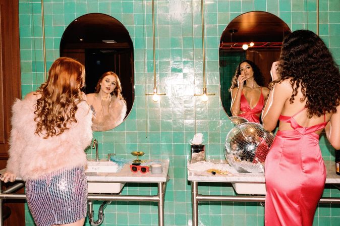 Two women getting ready at a retro green-tiled bathroom vanity: one in a pink feather jacket and sequin skirt touches up makeup in an oval mirror, the other in a satin pink dress applies lipstick beside a disco ball.