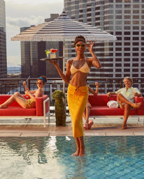 Sunset rooftop pool bar scene — sunglasses-clad server in a bikini top and yellow sarong balancing on the water while carrying a tray of colorful cocktails, guests lounging on red couches under a striped umbrella with a downtown skyscraper skyline in the background.