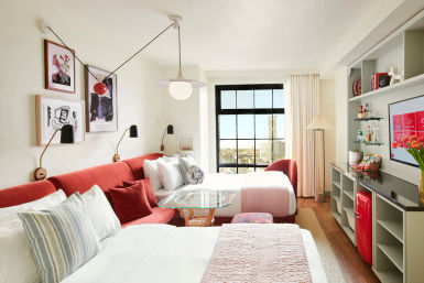 Bright, modern hotel room with large black-framed window showing an urban skyline, two beds with white linens and pink throws, red upholstered sofa, glass coffee table, wall art and reading lamps, built-in shelving with mini red fridge and snacks, flat-screen TV and floor lamp — cozy, cheery city stay.