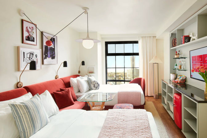 Bright, modern hotel room with large black-framed window showing an urban skyline, two beds with white linens and pink throws, red upholstered sofa, glass coffee table, wall art and reading lamps, built-in shelving with mini red fridge and snacks, flat-screen TV and floor lamp — cozy, cheery city stay.