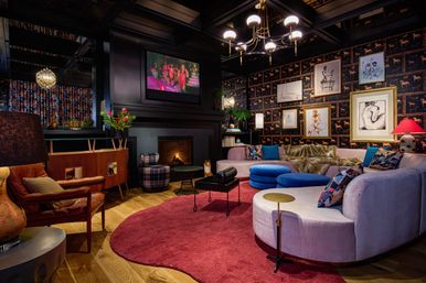 Eclectic cozy living room with curved blush sofa, blue round ottomans, red circular rug, fireplace with TV above, framed art gallery wall, dark patterned wallpaper and brass chandelier