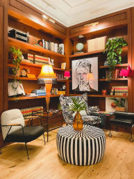 Cozy home library reading nook with wood-paneled built-in bookshelves, eclectic table and wall lamps, hanging plants and globe; patterned armchair, mid-century chair and black-and-white striped round ottoman with amber vase on hardwood floor.
