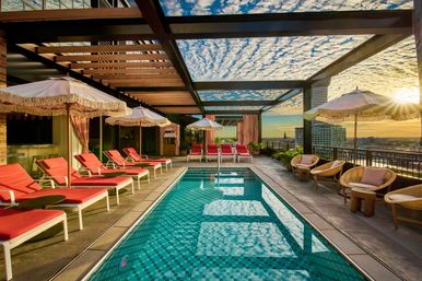 Rooftop downtown pool at sunset featuring a turquoise tiled lap pool, pink-cushioned lounge chairs and fringed umbrellas under a wooden pergola, with city skyline and golden sun on the horizon.