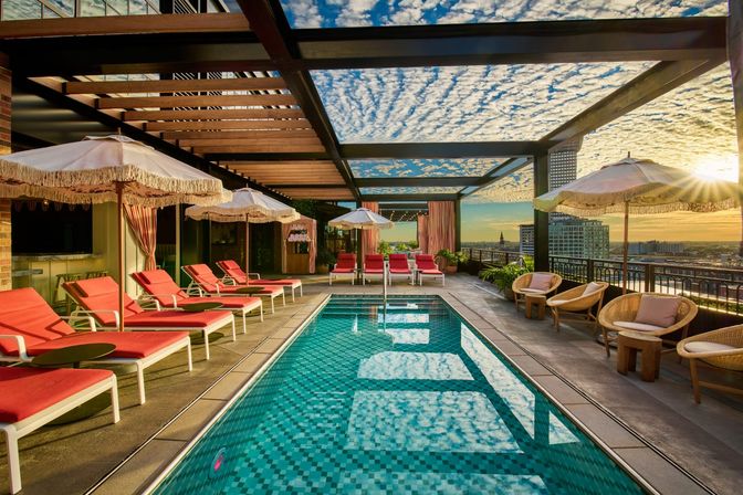 Rooftop downtown pool at sunset featuring a turquoise tiled lap pool, pink-cushioned lounge chairs and fringed umbrellas under a wooden pergola, with city skyline and golden sun on the horizon.