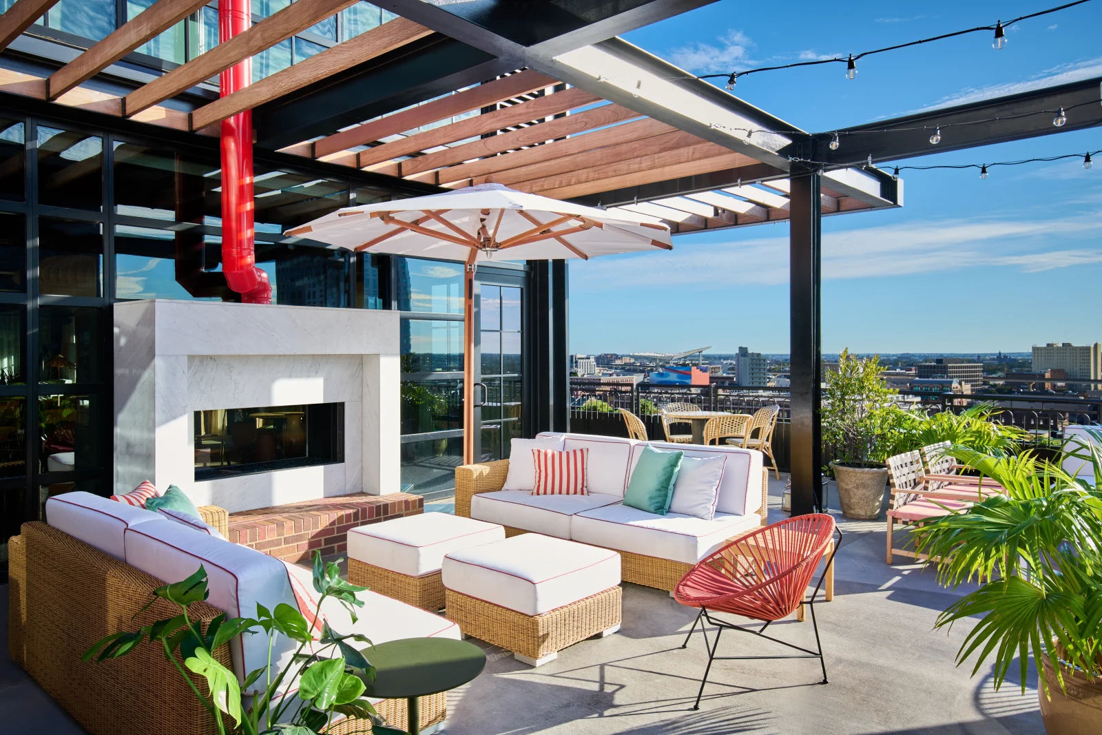 Sunny rooftop terrace lounge with wicker sofas, white cushions and ottomans, a large umbrella and modern marble fireplace under a wooden pergola, string lights, potted tropical plants and city skyline views.