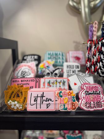 Colorful embroidered iron-on patches like “Let them” and novelty Moms Club designs displayed on a clear acrylic tray on a boutique store shelf.