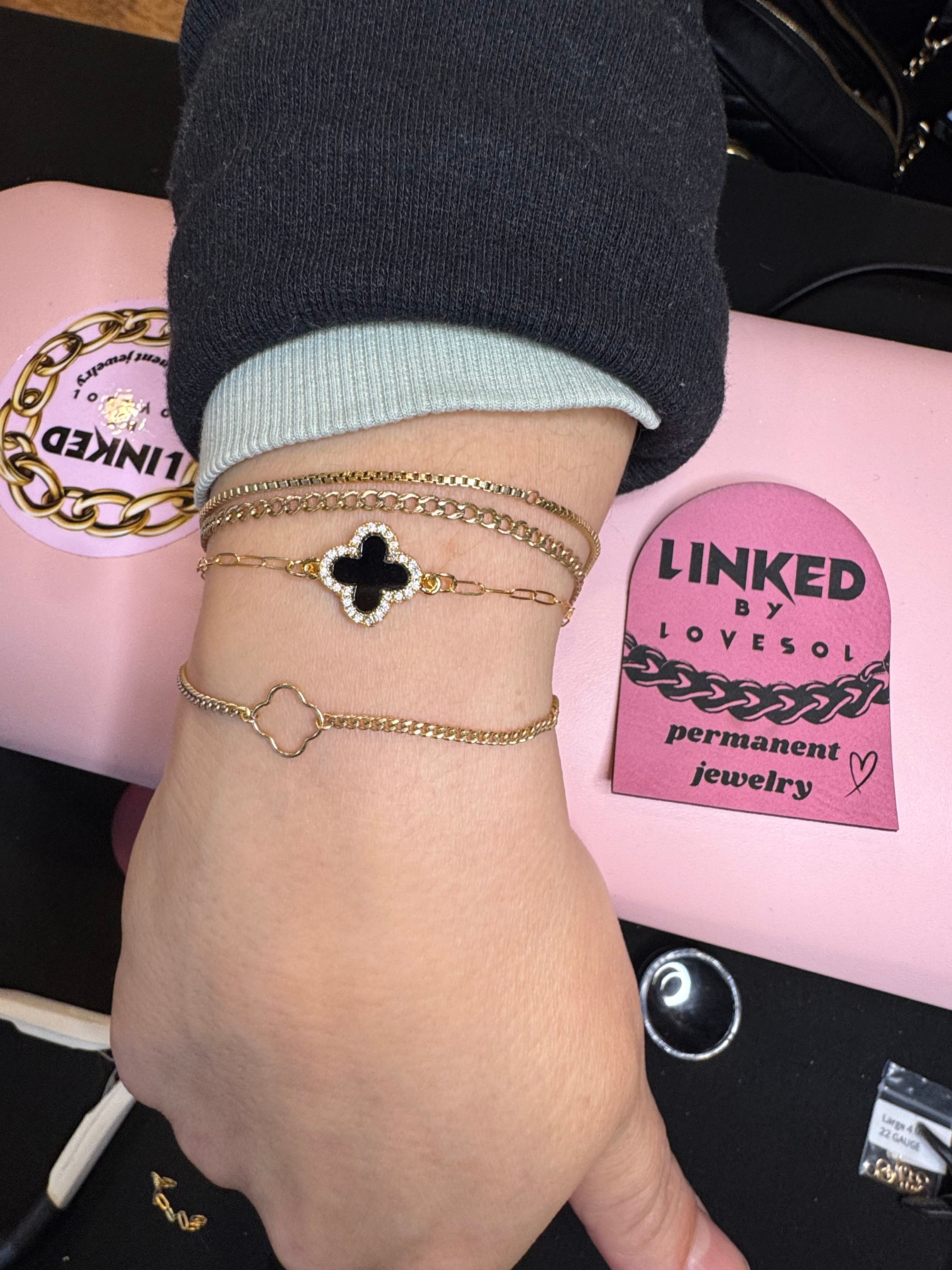 Wrist close-up wearing stacked gold bracelets with a black clover charm and delicate chains, photographed on a pink permanent jewelry studio mat