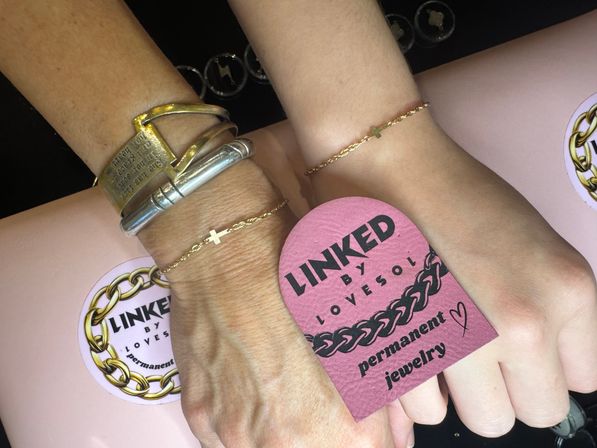 Close-up of two wrists wearing matching delicate gold cross chain bracelets; one wrist also has chunky silver and brass bangles, with a pink tag and stickers promoting permanent jewelry and chain designs on a pale pink background.