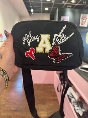 Black nylon crossbody bag held in a shop, decorated with embroidered patches reading 'girl gang', a gold-trimmed fuzzy letter A, a red butterfly and a skeleton-hand design.