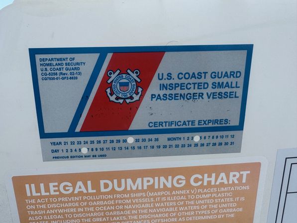 U.S. Coast Guard inspected small passenger vessel sticker with emblem and peel-out expiration calendar mounted above an 'Illegal Dumping' informational poster on a boat