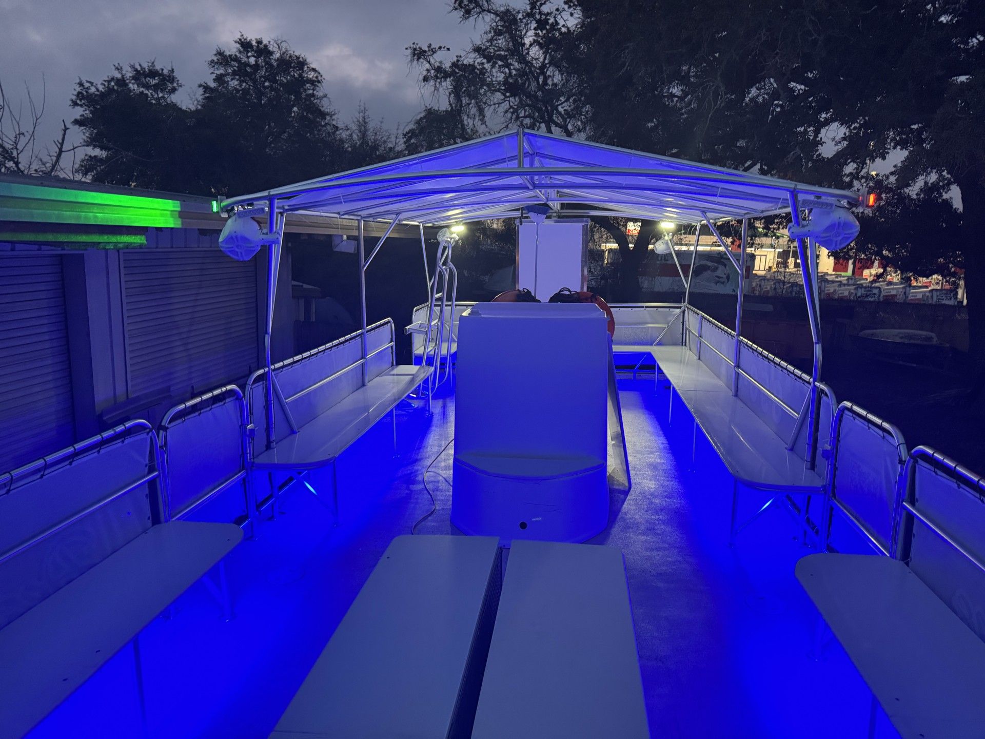 Covered pontoon boat deck at dusk with vibrant blue LED floor and canopy lighting, bench seating along both sides, life rings near the helm and trees and dock structures visible in the background.