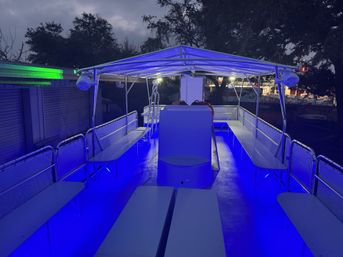 Covered pontoon boat deck at dusk with vibrant blue LED floor and canopy lighting, bench seating along both sides, life rings near the helm and trees and dock structures visible in the background.