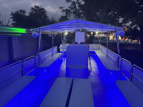 Covered pontoon boat deck at dusk with vibrant blue LED floor and canopy lighting, bench seating along both sides, life rings near the helm and trees and dock structures visible in the background.