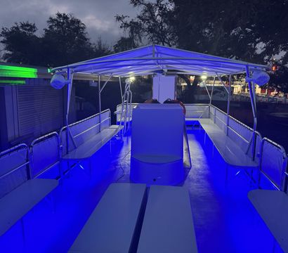 Covered pontoon boat deck at dusk with vibrant blue LED floor and canopy lighting, bench seating along both sides, life rings near the helm and trees and dock structures visible in the background.