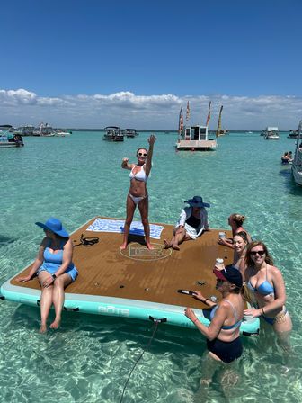 Women enjoying a sunny sandbar day on a floating dock in clear turquoise shallow water, surrounded by boats under a bright blue sky