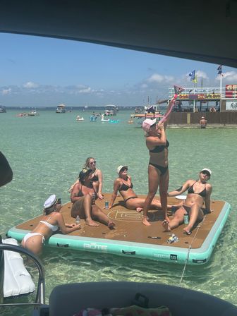 Sun-soaked beach scene with a group of women in bikinis relaxing and playing on a large floating dock in crystal-clear turquoise bay, surrounded by anchored boats and a lively waterfront shack with colorful flags under a bright blue sky.