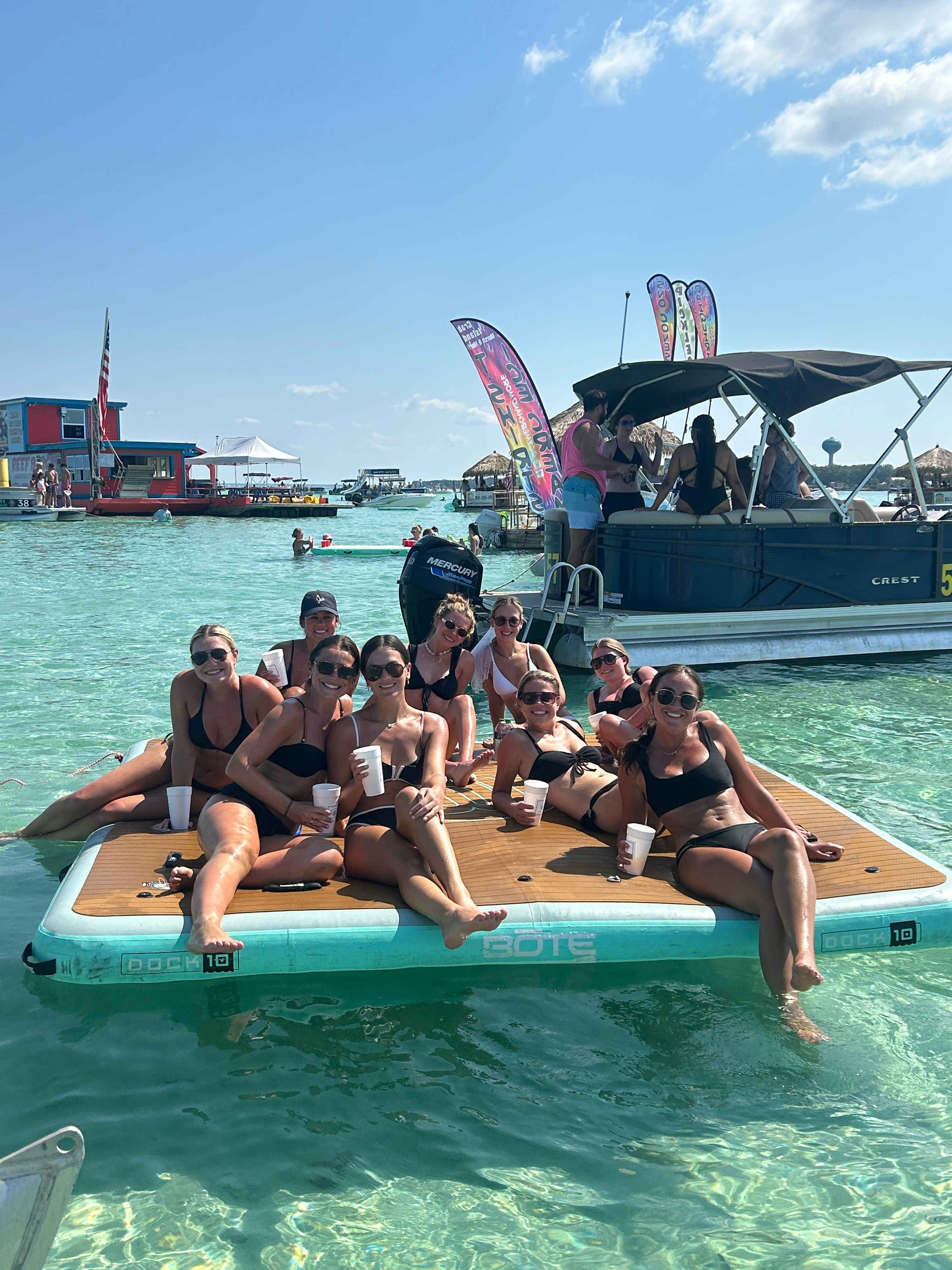 Smiling group of women in swimsuits lounging on a floating dock with drinks in clear turquoise shallow water, a nearby pontoon boat, colorful flags and sunny blue sky over a lively coastal lagoon.