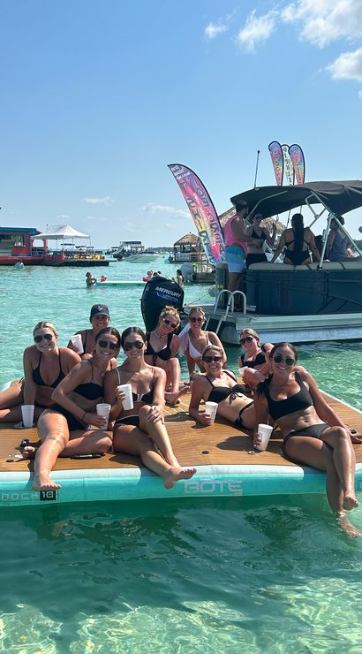 Smiling group of women in swimsuits lounging on a floating dock with drinks in clear turquoise shallow water, a nearby pontoon boat, colorful flags and sunny blue sky over a lively coastal lagoon.