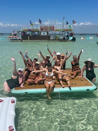 Group of women in swimsuits cheering and holding drinks on a floating dock in clear turquoise shallow water with a colorful floating general store and rental boat in the background under a sunny blue sky.