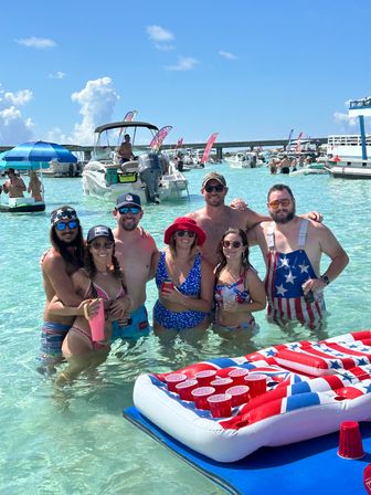 Group of friends posing waist-deep in clear turquoise shallow water at a busy sandbar, surrounded by anchored boats, colorful flags and inflatables including a red, white and blue beer-pong float under a bright sunny sky