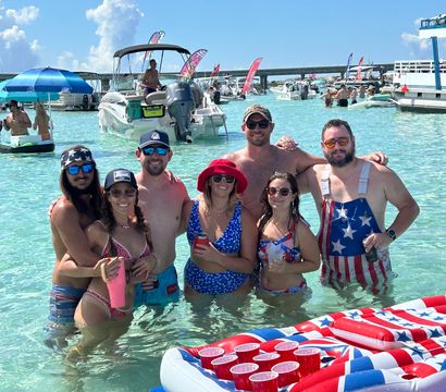Group of friends posing waist-deep in clear turquoise shallow water at a busy sandbar, surrounded by anchored boats, colorful flags and inflatables including a red, white and blue beer-pong float under a bright sunny sky