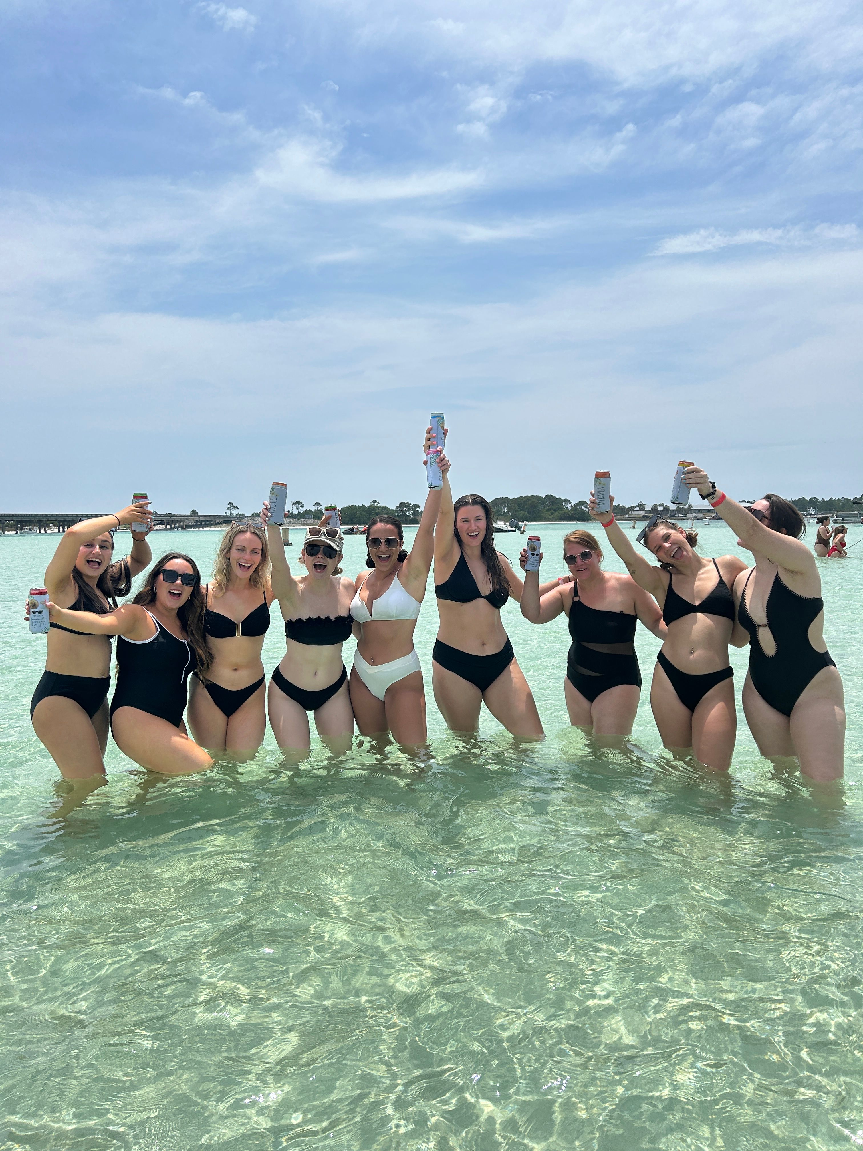 Group of friends in black and white bikinis cheering with canned drinks while standing in shallow turquoise beach water under a sunny blue sky
