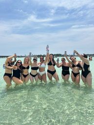 Group of friends in black and white bikinis cheering with canned drinks while standing in shallow turquoise beach water under a sunny blue sky