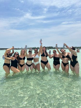 Group of friends in black and white bikinis cheering with canned drinks while standing in shallow turquoise beach water under a sunny blue sky