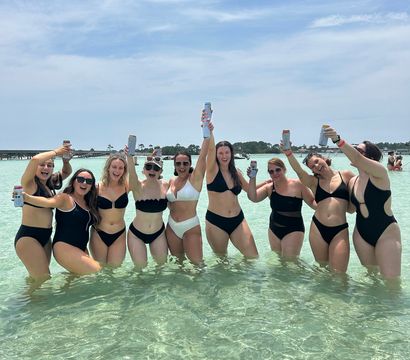Group of friends in black and white bikinis cheering with canned drinks while standing in shallow turquoise beach water under a sunny blue sky