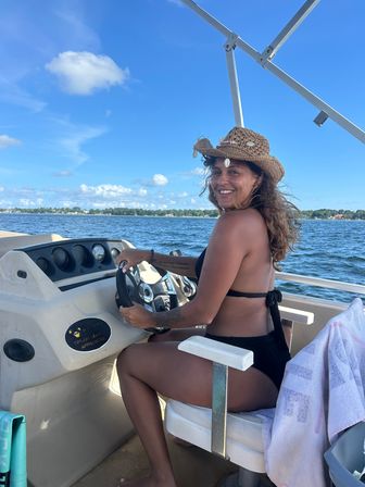 Smiling woman wearing a shell-decorated straw hat steering a boat on bright blue water with a sunny sky and distant shoreline