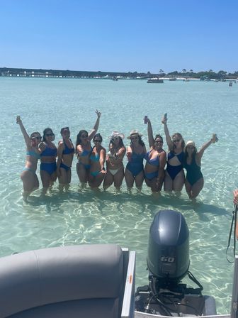 Group of women in swimsuits standing waist-deep in clear turquoise water, raising drinks and smiling with boats, a bridge and sandy shoreline in the sunny background.