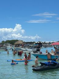 Crowded sandbar party with people wading in shallow turquoise water around anchored boats and a jet ski beneath a bright blue sky dotted with puffy clouds