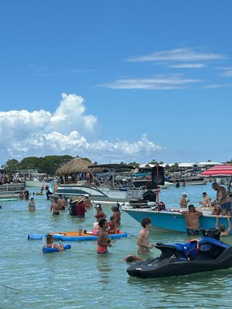 Crowded sandbar party with people wading in shallow turquoise water around anchored boats and a jet ski beneath a bright blue sky dotted with puffy clouds